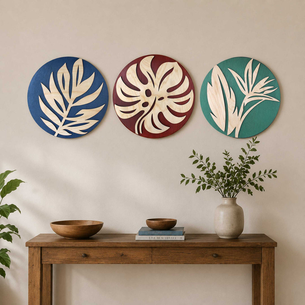 Tropical Botanical Wall Art Set of 3 circular wooden panels featuring cream cutout leaf silhouettes on blue, burgundy, and teal backgrounds, displayed above a wooden console table with plants and decor