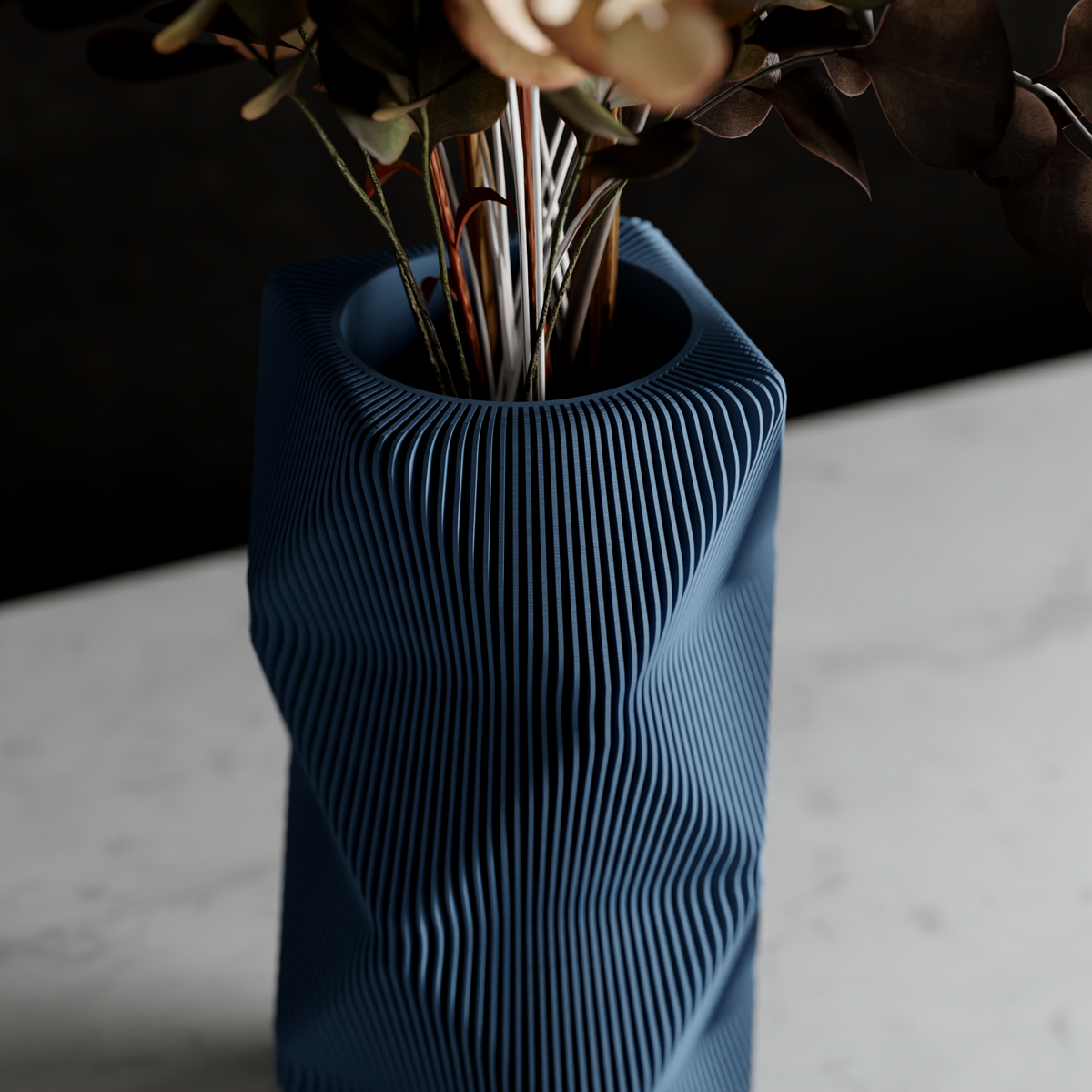 Close-up of Mountain Vase in deep slate blue with fine ridged topographic texture, holding dried botanical stems, displayed on a marble surface against a dark background
