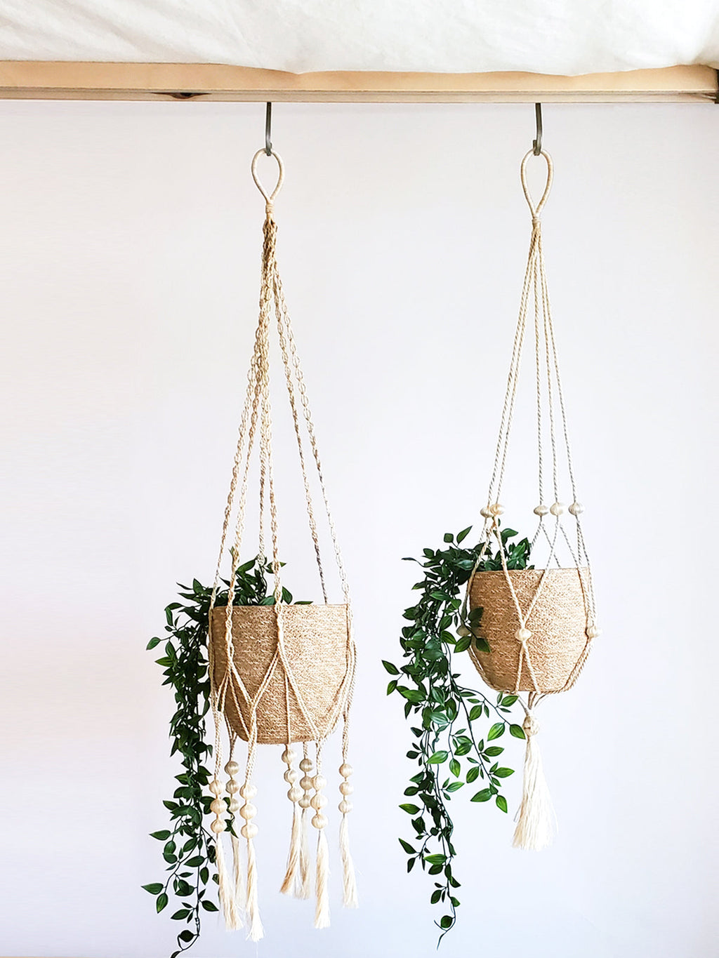 Two cream macrame plant hangers with wooden beads and tassel ends, each holding a woven seagrass basket with trailing green plants, hung from a wooden beam against a white wall