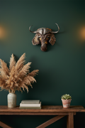A rustic metal African buffalo head wall mount displayed on a dark green wall above a wooden console table styled with pampas grass, books, and a small succulent.