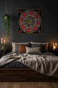 A dark bedroom scene with a bed dressed in gray and beige bedding, decorated with multiple pillows, beneath a large black wall tapestry featuring a colorful circular mandala design. The room has dark walls, wooden flooring, candles on bedside tables, and a hanging green plant.